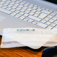 The top view of the workspace during the covid19 (coronavirus) is spreading with a laptop computer, nose mask, glasses, and a cup of herbal drink on a gray wooden table.The idea for coping and virus