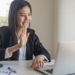 Happy young asian business woman waving hands to greeting partner during making video conference with her team. Happy young asian business woman waving hands to greeting partner during making video conference with her team.