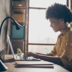 Profile side photo of smart cool afro american girl entrepreneur sit table use laptop work presentation in office loft workplace